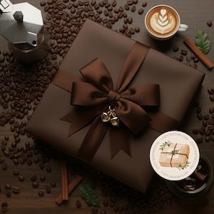 Puede incluir: Una caja de regalo marrón con una cinta y lazo a juego, adornada con pequeñas campanas doradas. La escena incluye granos de café, una taza de latte art, una cafetera italiana y palitos de canela, creando un ambiente de café.