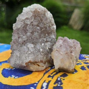 May include: Two raw amethyst crystal clusters with jagged, translucent purple and white points. The larger cluster is tall and upright, while the smaller cluster sits beside it, both displaying natural, unpolished mineral formations.