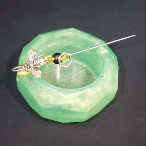 May include: A green, geometric-shaped dish with a silver bee charm and a silver and gold beaded chain. The dish is on a black surface.