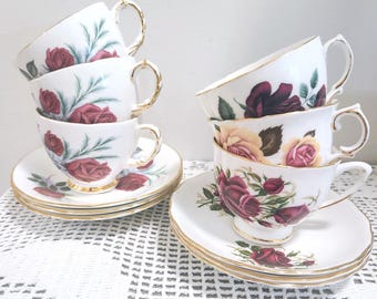 May include: A collection of white porcelain teacups and saucers featuring floral patterns of red and yellow roses with green leaves. Each piece is accented with a gold-trimmed rim and handle, arranged in two stacks on a white crocheted surface.