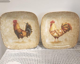 May include: Two square ceramic plates featuring a beige and tan mottled background with a decorative scroll border. Each plate displays a detailed illustration of a rooster with red, gold, and dark brown plumage standing in the center of the dish.