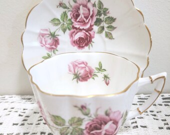 May include: A white porcelain teacup and saucer set featuring a scalloped edge with gold trim. The set is decorated with a pattern of pink roses and green leaves. The teacup has a single handle with gold accents and a floral motif on the interior.