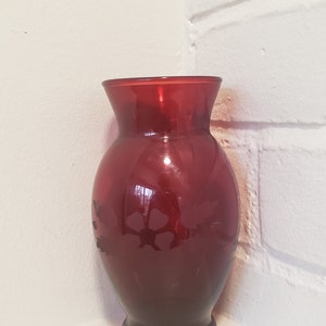 May include: A deep red glass vase with a floral design etched into the side. The vase has a rounded body and a flared lip.