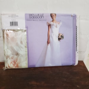 May include: A white wedding dress pattern with a purple background. The pattern features a model wearing a white wedding dress with a ruffled neckline and a bouquet of flowers. The text "Belleville Sassoom" and "Vogue Bridal Original" is printed on the pattern.