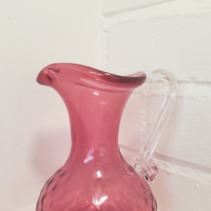 May include: A small, pink glass jug with a clear glass handle. The jug has a textured, dimpled surface.