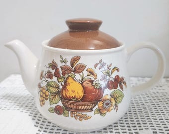 May include: A white ceramic teapot featuring a brown lid and a colorful fruit basket illustration on the front. The design includes a yellow pear, a red apple, and various green leaves and flowers, all centered on the rounded body of the teapot.
