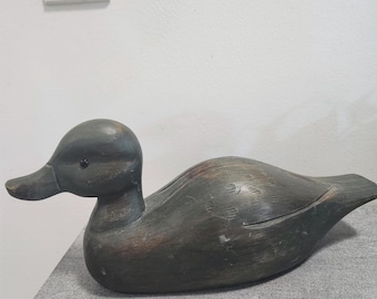 May include: A dark gray, carved wooden duck decoy with a smooth, rounded body and a flat tail. The surface features subtle, weathered wood grain textures and a small, dark, circular eye detail on the side of the head, facing toward the left.