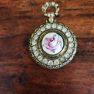 May include: A gold-toned pendant with a central circular design featuring a painted pink rose. The pendant is surrounded by clear crystals. The frame has an ornate design. A small loop is at the top for hanging, suitable for a necklace.