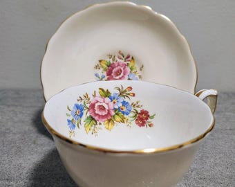 May include: Cream-colored teacup and saucer set with a floral design featuring pink, blue, and red flowers. The cup and saucer have gold trim along the edges. The saucer is scalloped.