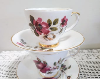 May include: Two white porcelain teacups and saucers stacked vertically. Each piece features a delicate floral pattern with deep pink and red roses, green leaves, and gold-trimmed scalloped edges. The top cup has a gold-painted base rim.