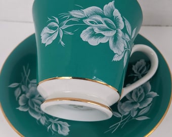 Può includere: Un set da tè composto da una tazza e un piattino color turchese con un motivo floreale bianco. La tazza e il piattino hanno un bordo dorato.