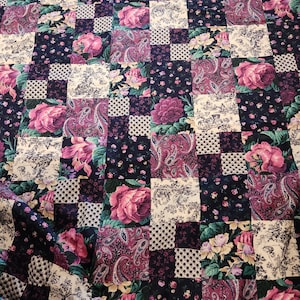 May include: A patchwork quilt with a mix of floral and paisley designs. The quilt uses squares in navy, purple, pink, and cream. Large pink roses and paisley patterns are featured, giving a vintage look. The quilt is made of fabric.