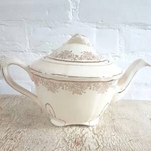 May include: A cream-coloured ceramic teapot featuring a delicate metallic gold floral pattern around the lid and the upper body. The teapot has a curved handle, a pointed spout, and a matching lid with a small decorative finial on top.