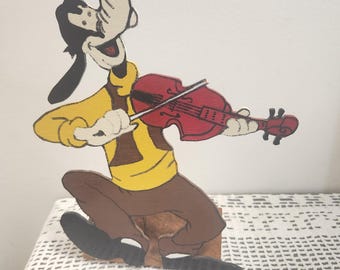 May include: A flat, cutout wooden figure of a cartoon dog character with black ears, a yellow shirt, and brown pants, depicted in a seated position while playing a red violin with a bow. The figure features bold black outlines and painted details.