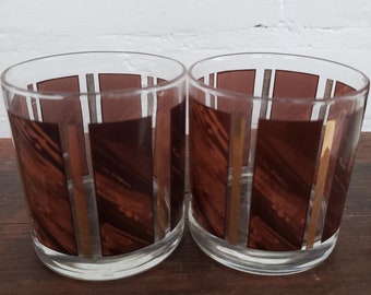 May include: Two clear glass tumblers with brown and gold striped designs. The glasses are empty and sitting on a wooden surface.