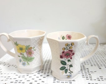 May include: Two off-white ceramic mugs with an octagonal shape and a textured basket-weave pattern on the exterior. Each mug features a colorful floral decal with yellow, pink, and green botanical details on the front and inside the rim.