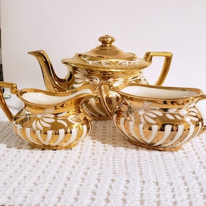 May include: A gold tea set with white floral designs. The teapot, creamer, and sugar bowl are all decorated with a gold and white striped pattern.