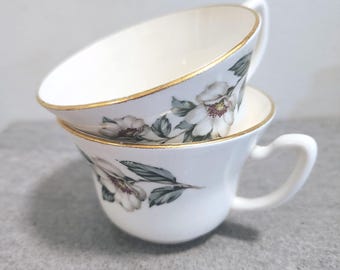 May include: Two stacked white ceramic teacups featuring a delicate floral pattern with white petals, dark centers, and muted green leaves. Each cup has a smooth, rounded handle and a thin, metallic gold rim along the top edge of the vessel.