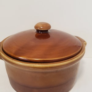 May include: A brown ceramic casserole dish with a lid. The dish has a rounded shape and a smooth, glossy finish.