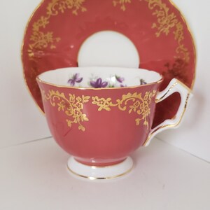 May include: A red teacup and saucer with a gold floral design and purple flowers. The saucer has a white centre.