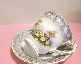 May include: A white teacup and saucer set with a floral design. The teacup features a gold handle and rim, and the saucer has a matching gold trim. The floral pattern includes yellow, purple, and pink flowers.