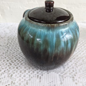 Op de afbeelding: Een bruine en blauwe geglazuurde keramische suikerpot met deksel. De pot is rond en heeft een gladde, glanzende afwerking.