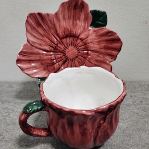 May include: A ceramic teacup and saucer set. The cup and saucer are shaped like a red flower with green leaves. The inside of the cup is white. The set is designed to look like a single flower.