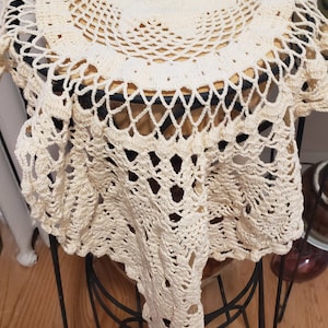 May include: A white crocheted doily with a delicate pattern, draped over a black metal plant stand. The doily is made of fine yarn and has a circular shape.