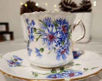May include: A white teacup and saucer with a blue and white floral pattern. The cup has a gold rim and the saucer has a gold band around the edge.
