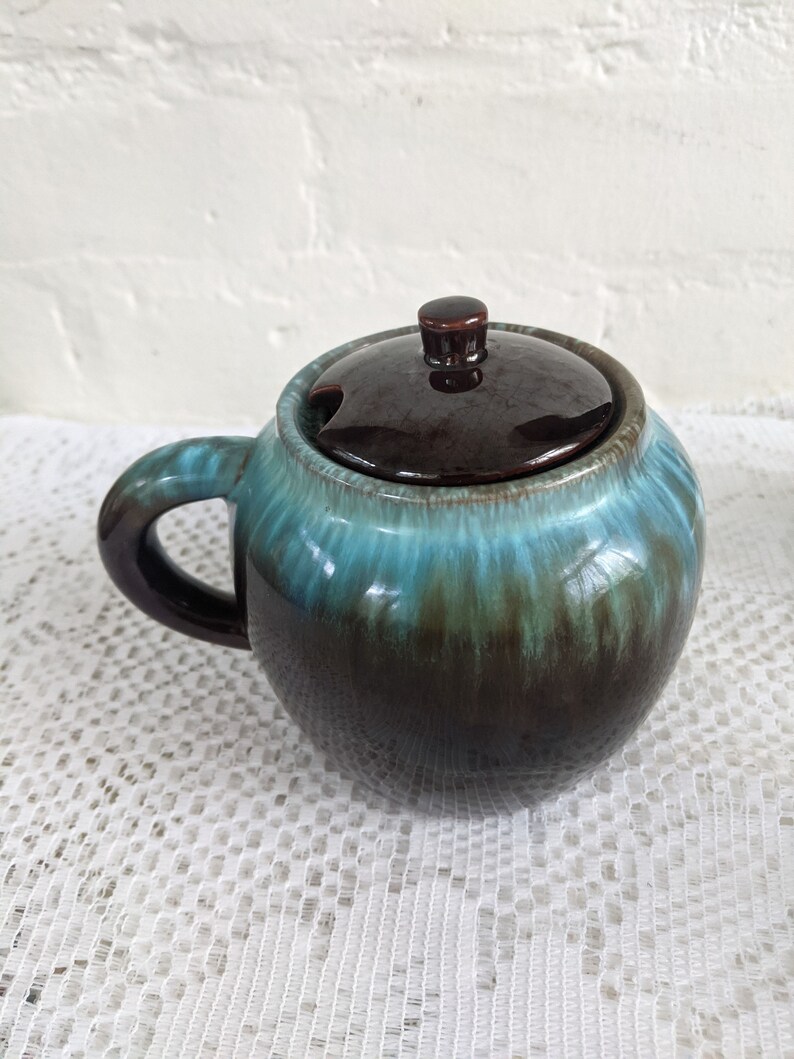 Op de afbeelding: Een bruine en blauwe geglazuurde keramische suikerpot met een deksel en een handvat. De kom heeft een gemarmerde blauwe en groene glazuur met bruine accenten.
