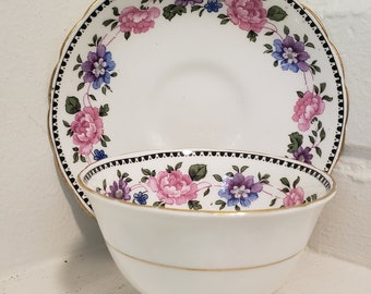 May include: A white teacup and saucer with a floral pattern in shades of pink, purple, and blue. The floral design features a repeating pattern of flowers and leaves around the rim of the saucer and teacup.