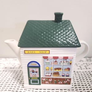 May include: A ceramic teapot shaped like a bake shop with a green shingled roof lid and a chimney. The white body features a green door, a large window displaying various cakes and pastries, and a sign above the window that reads BAKE SHOP.