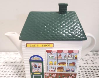 May include: A ceramic teapot shaped like a bake shop with a green shingled roof lid and a chimney. The white body features a green door, a large window displaying various cakes and pastries, and a sign above the window that reads BAKE SHOP.