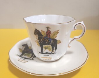 May include: White teacup and saucer set with gold trim. The teacup and saucer feature a design of a mounted RCMP officer and the text "R.C.M.P. Niagara Falls". The teacup has a curved handle.