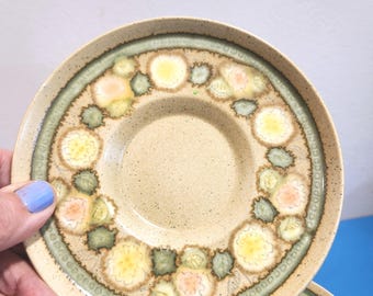 May include: Two beige ceramic saucers with a floral design. The saucers have a speckled texture and a green border. The floral pattern includes yellow, peach, and green elements. The saucers are stacked, with one partially visible.