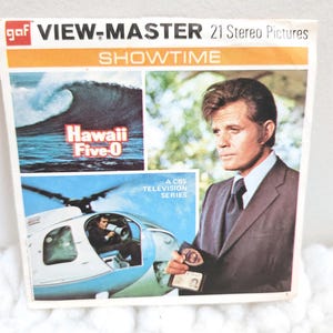 May include: A vintage View-Master Showtime packet featuring images from the television series Hawaii Five-0. The packet has a white, orange, and blue color scheme, with images of a wave, a helicopter, and a man in a suit.