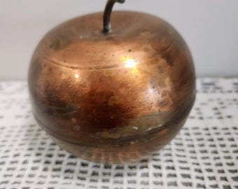 May include: A spherical copper apple sculpture with a polished metallic finish and a dark, curved stem. The surface displays natural variations in color, ranging from bright copper tones to darker, weathered patches across the round body.