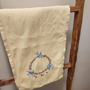 Peut inclure: Une serviette de thé en lin crème avec un motif brodé à la main d'oiseaux bleus dans une couronne de fleurs roses.