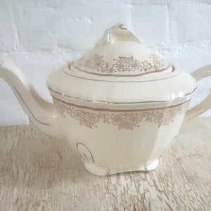 May include: A white teapot with a gold floral design. The teapot has a lid and a spout. The teapot is sitting on a wooden surface.