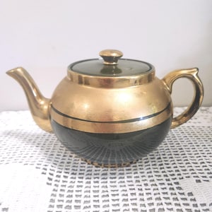 May include: A round teapot featuring a metallic gold upper section and a solid black lower section, separated by a thin black horizontal band. The lid is dark green with a gold knob, and the teapot includes a curved spout and a handle.