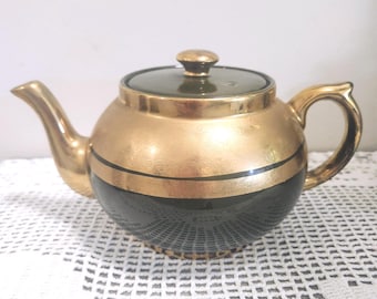 May include: A round teapot featuring a metallic gold upper section and a solid black lower section, separated by a thin black horizontal band. The lid is dark green with a gold knob, and the teapot includes a curved spout and a handle.