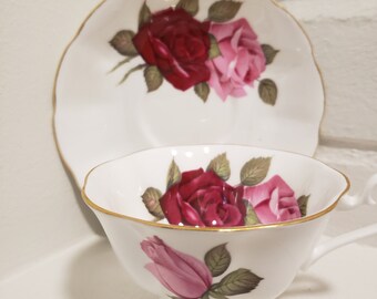 May include: A white teacup and saucer with a gold rim. The teacup and saucer feature a floral design with pink and red roses.