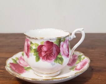 May include: A white teacup and saucer with a pink rose floral pattern. The cup has a gold rim and the saucer has a scalloped edge.