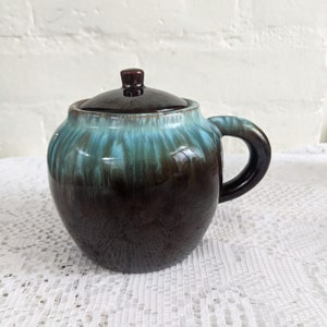 Op de afbeelding: Een bruine en teal geglazuurde keramische pot met een deksel. De pot heeft een rond lichaam en een handvat. De glazuur heeft een gemarmerd effect.