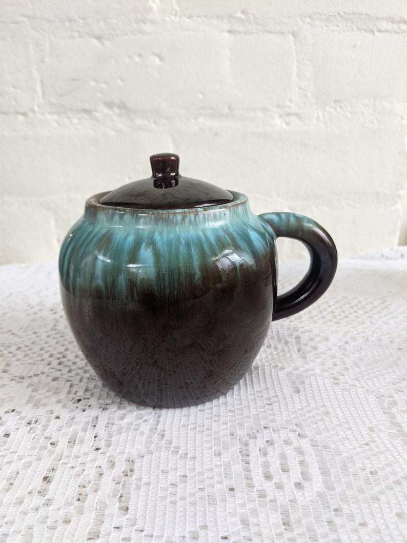 Op de afbeelding: Een bruine en teal geglazuurde keramische pot met een deksel. De pot heeft een rond lichaam en een handvat. De glazuur heeft een gemarmerd effect.