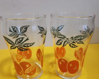 May include: Two clear drinking glasses with a gold rim. The glasses are decorated with orange fruit and green leaf designs. The fruit appears to be oranges. The glasses are on a yellow surface.