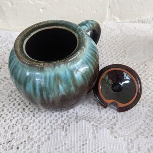 Op de afbeelding: Een bruine en teal geglazuurde keramische suikerpot met een deksel. De pot heeft een ronde vorm en een handvat aan de zijkant. Het deksel heeft een klein tuitje om te gieten.