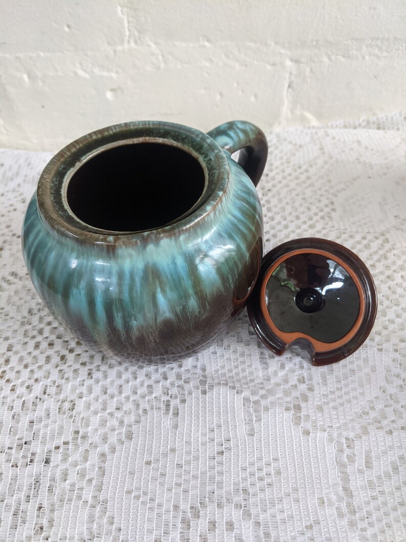 Op de afbeelding: Een bruine en teal geglazuurde keramische suikerpot met een deksel. De pot heeft een ronde vorm en een handvat aan de zijkant. Het deksel heeft een klein tuitje om te gieten.