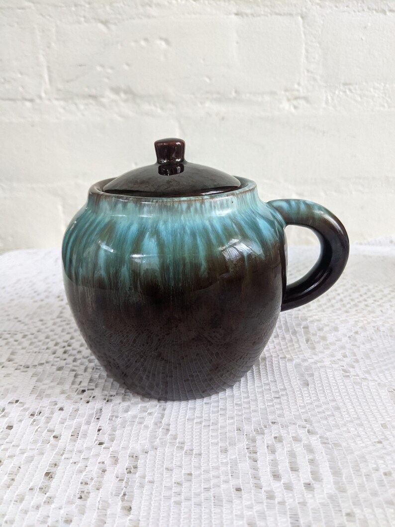 Op de afbeelding: Een bruine en turquoise geglazuurde keramische roomkan met een deksel en een handvat. De roomkan heeft een gemarmerde glazuur met turquoise en bruine strepen.