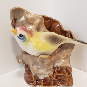 May include: A ceramic planter shaped like a tree stump with a yellow and white bird perched on a branch. The bird has blue and red accents on its head.
